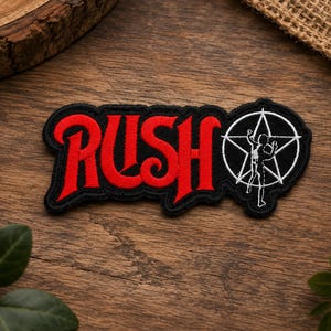 May include: An embroidered patch featuring the word RUSH in bold red lettering with a black border. To the right of the text is a white circular emblem containing a white five-pointed star and a white silhouette of a person standing inside.