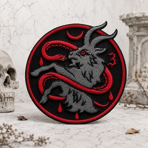 May include: A circular embroidered patch featuring a grey goat with red eyes, surrounded by a red snake on a black background. The design includes a red crescent moon, three red teardrop shapes, and a red number three, all enclosed by a red border.