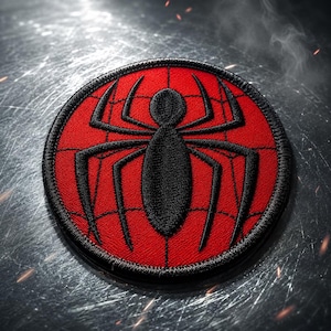 May include: A circular embroidered patch featuring a vibrant red background with a black spider emblem at the center. The design includes a thin black web pattern stitched over the red surface, finished with a thick black stitched border edge.