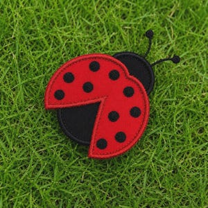 Puede incluir: Un parche bordado con forma de mariquita que presenta un diseño de alas de color rojo brillante con puntos circulares negros. El parche tiene una base negra, una sección de cabeza negra y dos antenas negras finas en la parte superior.