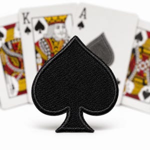 May include: A black embroidered patch in the shape of a spade suit symbol. The patch features a dense, textured thread construction with a clean, stitched border, presented against a background of blurred playing cards including a king and an ace.