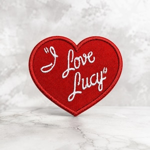 May include: A heart-shaped embroidered patch featuring a vibrant red fabric base with a dark red stitched border. The text "I Love Lucy" is embroidered in white cursive script across the center of the heart, creating a clear and bold contrast.