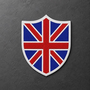 Può includere: Una toppa ricamata a forma di scudo con il design della bandiera Union Jack. La toppa presenta una croce rossa centrale e strisce diagonali rosse su sfondo blu, il tutto delineato da un bordo bianco spesso e dettagli di cucitura bianchi.