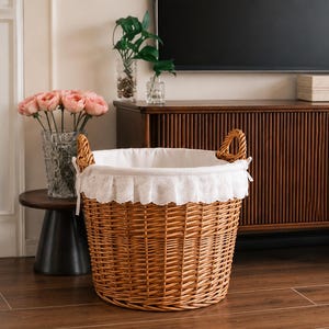 May include: A round, natural-coloured woven wicker basket featuring two sturdy side handles and a removable white fabric liner. The liner is finished with a decorative white eyelet lace trim that folds over the top edge of the basket.