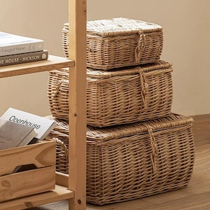 May include: A stack of three rectangular light brown wicker storage baskets with lids. Each basket features a woven texture and a braided loop closure at the center front. The baskets are arranged in a vertical column, decreasing in size from bottom to top.