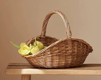 May include: A light brown woven wicker basket with a curved rim and a single arched handle. The basket features a natural wood texture and contains a small cluster of pale yellow tulip flowers tucked into one side of the open interior space.