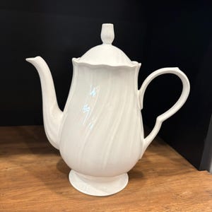 May include: A white porcelain teapot featuring a glossy finish, a curved spout, a C-shaped handle, and a removable lid with a finial. The body of the teapot displays a subtle, vertical spiral-ribbed texture and a scalloped rim edge.