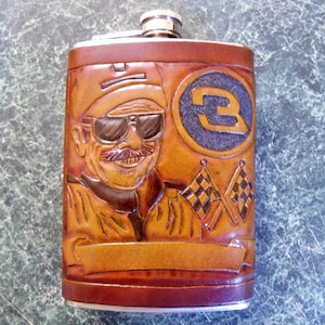 May include: A brown leather flask with a carved image of a race car driver wearing sunglasses and a cap. The flask has a silver top and a checkered flag design. The number "3" is carved into a circle on the flask.