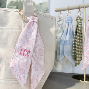 May include: A cream-colored canvas tote bag featuring a pink and white floral patterned fabric ribbon tied to the handle with the embroidered monogram LCC. Additional fabric ribbons in various patterns hang from a gold metal display stand nearby.