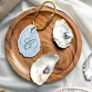 May include: A light blue embroidered oyster-shaped keychain featuring a dark gray cursive letter C, attached to a gold-toned metal carabiner. The keychain is displayed alongside two natural oyster shells with gold-painted edges on a round wooden tray.