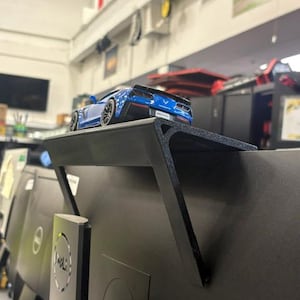 May include: A black plastic monitor top shelf mounted on the upper edge of a computer screen. A small blue toy sports car is positioned on the flat surface of the shelf. The shelf features a triangular support bracket extending down the back.