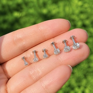 May include: A set of six silver-toned labret studs, each featuring a clear, round-cut gemstone set in a four-prong basket. The studs are arranged in a row, showcasing varying post lengths and consistent circular stone settings with metallic bases.