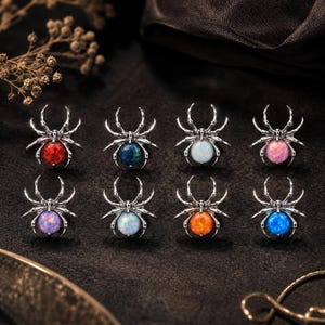Può includere: Otto spille o broche a forma di ragno color argento, ognuna con una pietra cabochon rotonda e iridescente di colore diverso, tra cui rosso, blu scuro, bianco, rosa, viola, azzurro, arancione e blu brillante, disposte su due file.