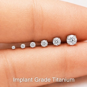 May include: A row of six round, clear, faceted stud earrings in graduated sizes, set in silver-toned metal. The text Implant Grade Titanium is displayed at the bottom. Each earring features a central stone held by small metal prongs.