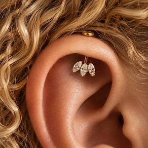 May include: A gold-toned ear piercing stud featuring three marquise-cut clear stones arranged in a fan-like cluster. The jewelry is secured with a small, round gold-toned backing, positioned within the upper cartilage area of the ear.