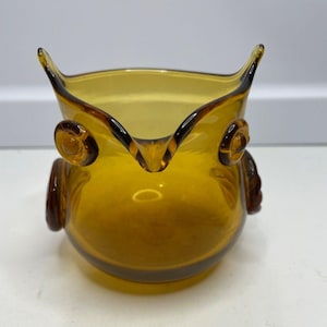 Pode incluir: Uma leiteira de vidro âmbar translúcido em forma de coruja. O recipiente apresenta dois olhos circulares em relevo, protuberâncias pontiagudas semelhantes a orelhas na borda superior e detalhes de asas moldados nas laterais do corpo.