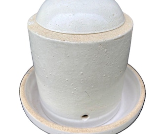 May include: A cylindrical ceramic butter keeper with a textured off-white glaze and a matching lid. The base features a wide circular tray with a small drainage hole near the bottom edge. The exterior surface has a rustic, speckled appearance.