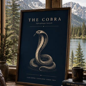 Op de afbeelding: Een ingelijste verticale poster met een donkerblauwe achtergrond en een gedetailleerde illustratie van een cobra. De tekst luidt THE COBRA, Ophiophagus hannah en CALM POWER. CLEAR MIND. STRIKE TRUE. De poster zit in een houten lijst.