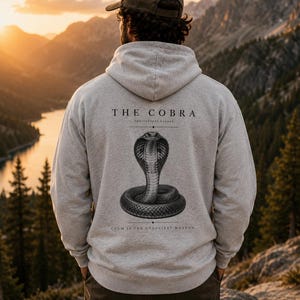 Könnte beinhalten: Ein hellgrauer Kapuzenpullover mit einem schwarzen Grafikdruck auf der Rückseite. Das Design zeigt eine zusammengerollte Kobra mit dem Text THE COBRA, Ophiophagus hannah und dem Satz CALM IS THE DEADLIEST WEAPON in einer klaren, dunklen Schrift.