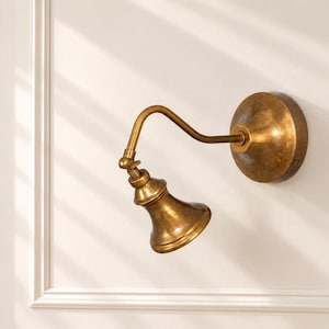 Può includere: Una lampada da parete con finitura in ottone anticato, dotata di una piastra di base circolare, un braccio curvo regolabile e un paralume a forma di campana. L'apparecchio in metallo presenta una superficie usurata e un giunto orientabile.