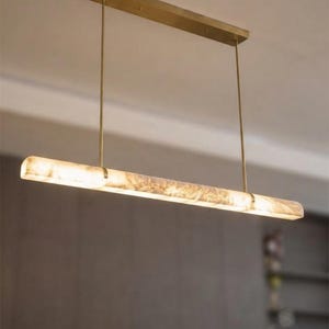 Könnte beinhalten: Eine lineare Pendelleuchte mit einem langen, zylindrischen, lichtdurchlässigen Steinschirm mit marmorierter Textur. Die Leuchte ist an einer rechteckigen Messing-Deckenhalterung mit zwei dünnen, vertikalen Messingstangen befestigt.