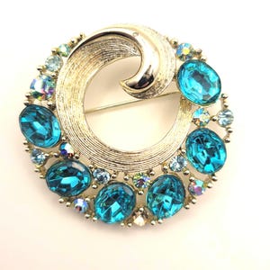 May include: A circular gold-tone metal brooch featuring a textured swirl design. The piece is accented with multiple oval-shaped bright blue faceted stones and smaller iridescent round crystals arranged along the outer edge of the frame.