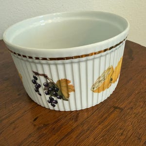 May include: A white ceramic ramekin with vertical fluted sides and a gold-toned rim. The exterior features a decorative pattern of dark purple berries on a stem with green leaves and a yellow fruit slice, set against a plain white background.