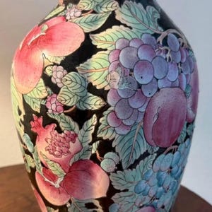 May include: A black ceramic vase featuring a vibrant, multi-colored botanical pattern of grapes, pomegranates, and various fruits with green leaves. The vase has a flared rim, a rounded body, and a narrow base, showcasing a detailed, colorful design.