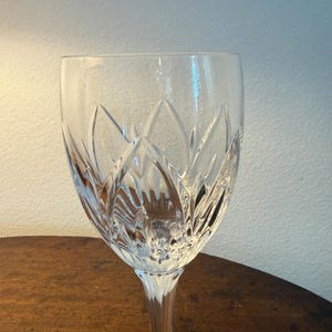 May include: A clear crystal wine glass featuring a deep, vertical leaf-like cut pattern around the bowl. The stem is smooth and leads down to a circular base, with the glass reflecting light through its intricate, faceted design.
