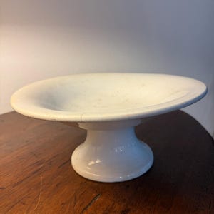 Puede incluir: Un cuenco de cerámica blanca sobre pedestal con una base ancha y poco profunda y una base acampanada. La superficie presenta una textura de esmalte craquelado sutil con manchas marrones tenues y una grieta visible en el interior.