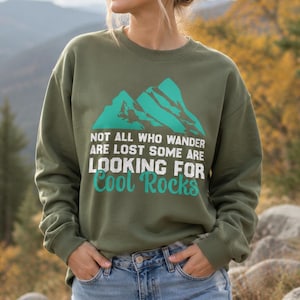 May include: An olive green crewneck sweatshirt featuring a teal mountain graphic and white and teal text that reads: Not all who wander are lost some are looking for cool rocks. The garment has long sleeves and a ribbed neckline and cuffs.