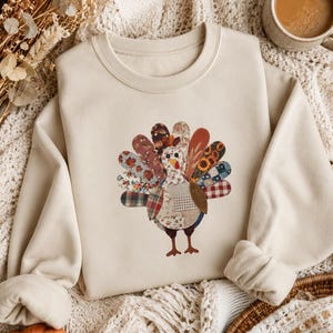 Peut inclure: Un sweat-shirt à col rond beige avec un motif central représentant une dinde composée de divers empiècements de style tissu. Les plumes de la queue de la dinde présentent des motifs variés, notamment floraux, à carreaux et tournesols.