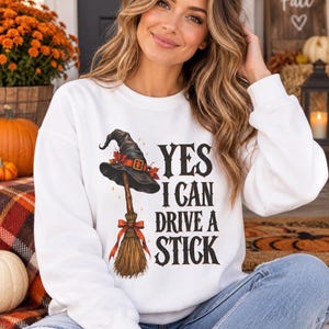 Op de afbeelding: Een wit sweatshirt met lange mouwen en een afbeelding van een zwarte heksenhoed op een houten bezemsteel met een oranje strik. De tekst "YES I CAN DRIVE A STICK" staat in een zwart lettertype. Het ontwerp bevat kleine oranje blaadjes.