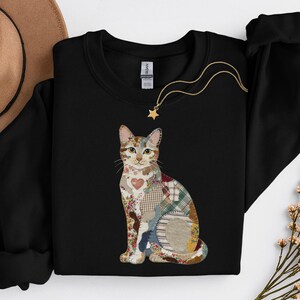 Può includere: Una felpa nera a maniche lunghe con una grafica centrale raffigurante un gatto seduto composto da vari motivi in tessuto colorato, inclusi disegni floreali, a quadri e a forma di cuore. Il design del gatto è realizzato in stile patchwork.