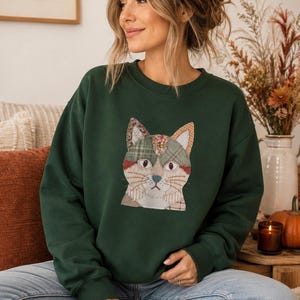 Puede incluir: Una sudadera de cuello redondo en color verde oscuro con un gráfico central de una cara de gato construida con varios parches de tela estampada. El diseño del gato incluye secciones de cuadros, flores y colores sólidos en tonos suaves.