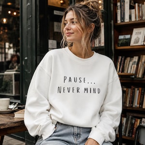 May include: A white long-sleeve crewneck sweatshirt featuring the black text PAUSE... NEVER MIND printed on the front. The garment has a relaxed fit with ribbed cuffs and a ribbed neckline, constructed from a solid-colored fabric.