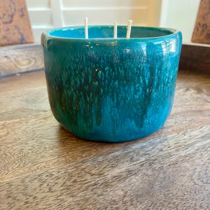 May include: A round ceramic candle vessel featuring a glossy, deep teal glaze with a dark, drippy, vertical pattern. The container has a smooth, curved exterior and contains three white cotton wicks extending from the top opening of the vessel.