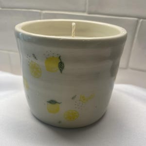 May include: A white ceramic candle vessel with a tapered cylindrical shape, featuring a repeating pattern of yellow lemons, green leaves, and small black dots. A single white wick is centered in the opening of the container.