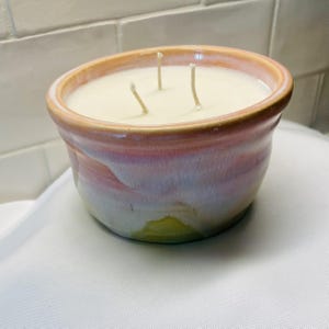 May include: A three-wick candle in a round, ceramic bowl with a glossy glaze featuring a blend of soft pink, lavender, and yellow hues. The wax is off-white and smooth, filling the bowl to the rim, with three cotton wicks protruding from the center.