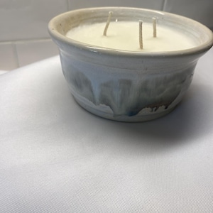 May include: A three-wick candle in a round, hand-thrown ceramic bowl. The bowl features a cream-colored upper section and a lower section with a reactive glaze in shades of blue, gray, and brown that drips down the sides of the vessel.