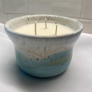 May include: A three-wick candle in a speckled ceramic bowl. The bowl features a white upper section and a light blue lower section with a tan band in the middle. The wax is off-white and fills the bowl to the rim, with three vertical wicks.