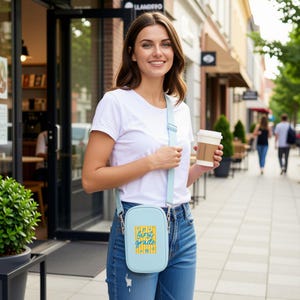 May include: A light blue crossbody bag with an adjustable strap, featuring a yellow vertical striped graphic and the text "first grade" in blue script. The bag is worn over a white t-shirt and blue jeans, with a coffee cup held in one hand.