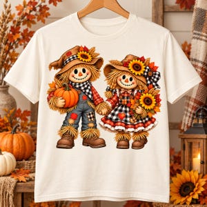 Peut inclure: Un t-shirt à manches courtes couleur crème avec une illustration de deux épouvantails se tenant la main. Ils portent des chapeaux ornés de tournesols, des écharpes à carreaux et des vêtements automnaux. L'un tient une citrouille.