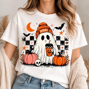 Peut inclure: Un t-shirt blanc avec un motif représentant un fantôme portant un bonnet orange sur lequel est écrit SPOOKY. Le fantôme tient un gobelet à café avec un visage de citrouille. Le design inclut deux citrouilles, un crâne, des chauves-souris, des étoiles et un croissant de lune sur un fond à damier noir et blanc.
