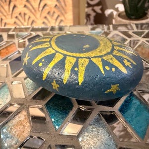 May include: A smooth, oval-shaped river stone painted in a deep navy blue color. The top surface features a hand-painted golden yellow sun design with radiating rays and several small, five-pointed stars scattered around the central sun motif.