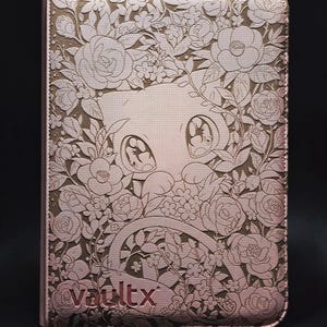 May include: A rose gold coloured binder with a textured surface featuring an embossed illustration of a cat-like creature surrounded by various flowers and leaves. The word vaultx is embossed in the bottom left corner of the rectangular cover.