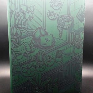 May include: A rectangular green binder featuring a debossed illustration of a creature resting on a counter surrounded by various potted plants, hanging greenery, and a window scene. The cover has a textured surface with dark green line art.