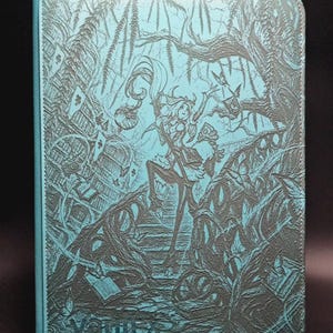 May include: A rectangular turquoise binder featuring a dark, intricate embossed illustration of a figure standing on a path surrounded by twisted trees, floating books, and foliage. The word Vaultx is embossed in the bottom left corner of the cover.