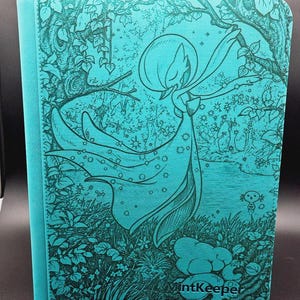 May include: A rectangular turquoise binder featuring a detailed dark teal line art illustration of a Gardevoir character standing in a forest setting with foliage and stars. The word MintKeeper is embossed in the bottom right corner of the cover.