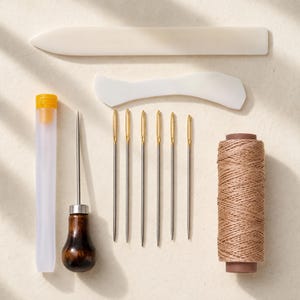 May include: A leather crafting tool set including a long white bone folder, a curved white creaser, a clear plastic needle case, an awl with a dark brown wooden handle, six silver needles with gold eyes, and a spool of light brown waxed thread.